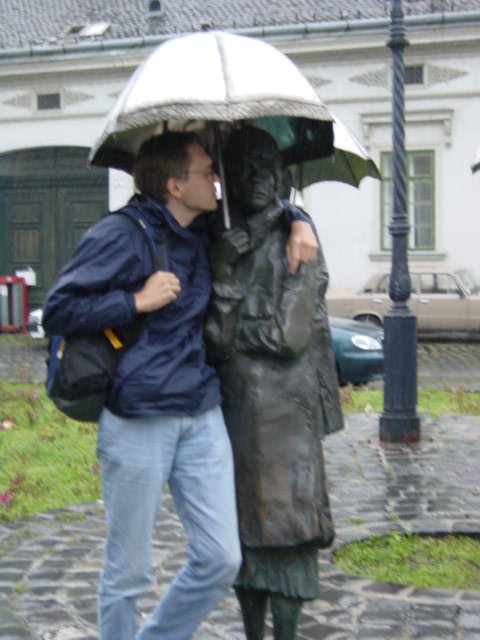Max standing near a statue, under the statue's umbrella with his arm on its shoulder and about to kiss its cheek