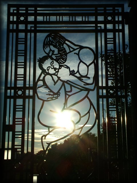 The sun setting slowly behind an iron gate featuring a man protectively cradling an infant. Vigeland Sculpture Park.