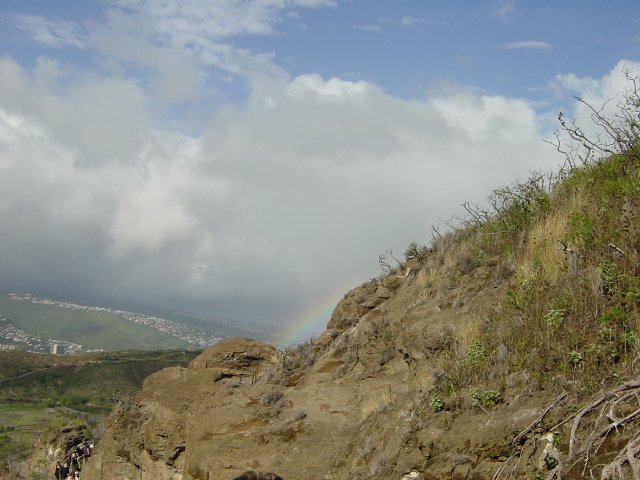 Small part of the rainbow hidden behind rocks, houses-covered hill, clouds and sky
