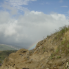 Small part of the rainbow hidden behind rocks, houses-covered hill, clouds and sky