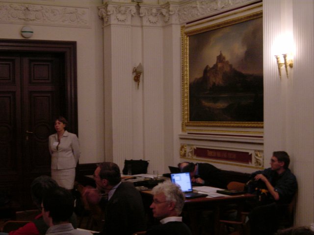 Marie standing by the door and laptop table, Vincent listening to a demo of his presentation, Max