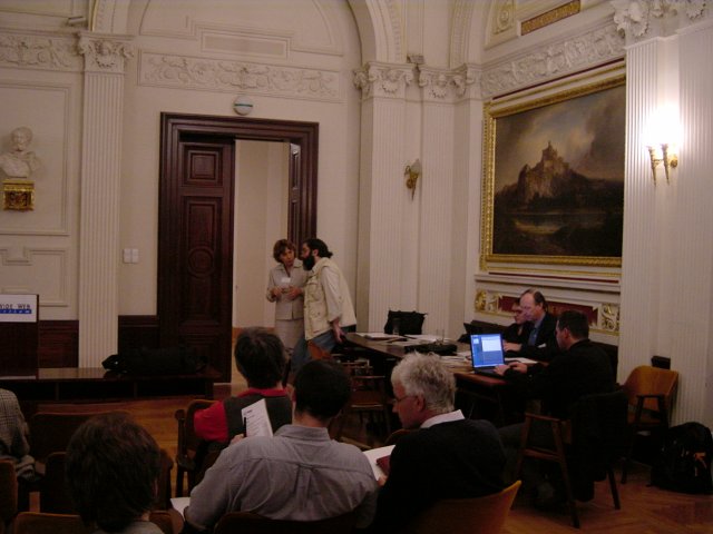 Maire and Ivan confering by the door, some attendees, Vincent and Max at the table